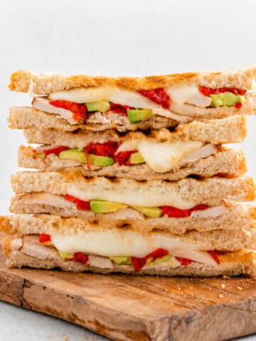 Four grilled sandwiches stacked on a wooden board, filled with cheese, turkey, avocado, and roasted red peppers.
