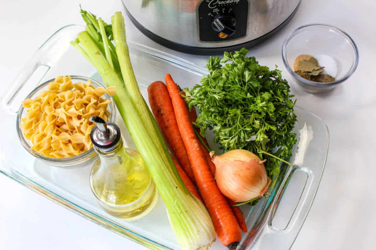 Ingredients for homemade Panera chicken noodle soup arranged beside a slow cooker.