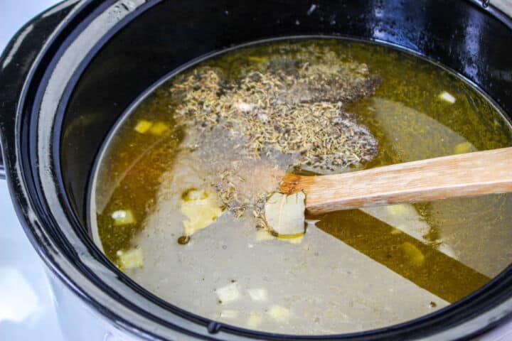 Broth, vegetables, thyme, and bay leaves stirred in a slow cooker for chicken noodle soup.