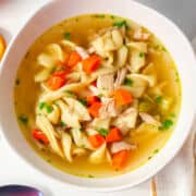 Chicken noodle soup with chicken pieces, egg noodles, carrots, and celery in clear broth, topped with chopped parsley.