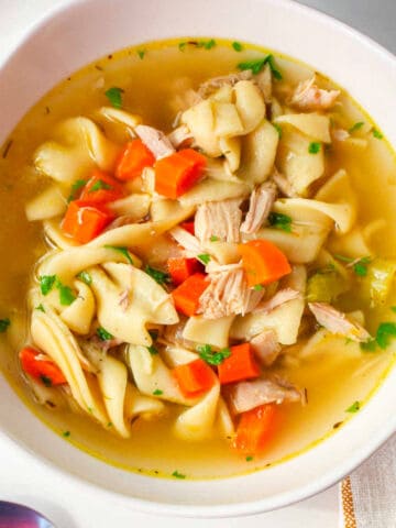 Chicken noodle soup with chicken pieces, egg noodles, carrots, and celery in clear broth, topped with chopped parsley.