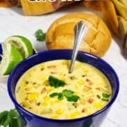 Blue bowl of Panera corn chowder with cilantro, spoon inside. Bread roll, lime, cilantro, and a colorful towel nearby.