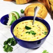 Blue bowl of Panera summer corn chowder with cilantro garnish, spoon, lime half, bread roll, and cilantro on white surface.
