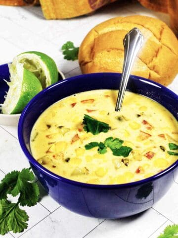 Blue bowl of Panera summer corn chowder with cilantro garnish, spoon, lime half, bread roll, and cilantro on white surface.