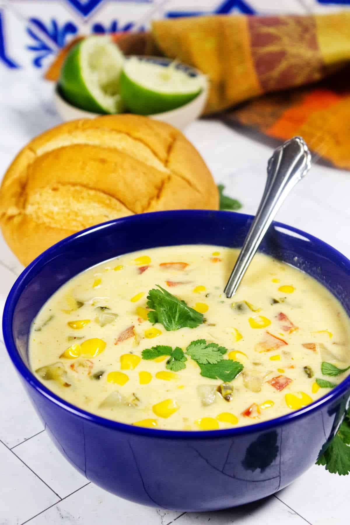 Blue bowl of Panera Summer Corn Chowder with cilantro, bread roll, lime wedges, spoon, and colorful napkin.