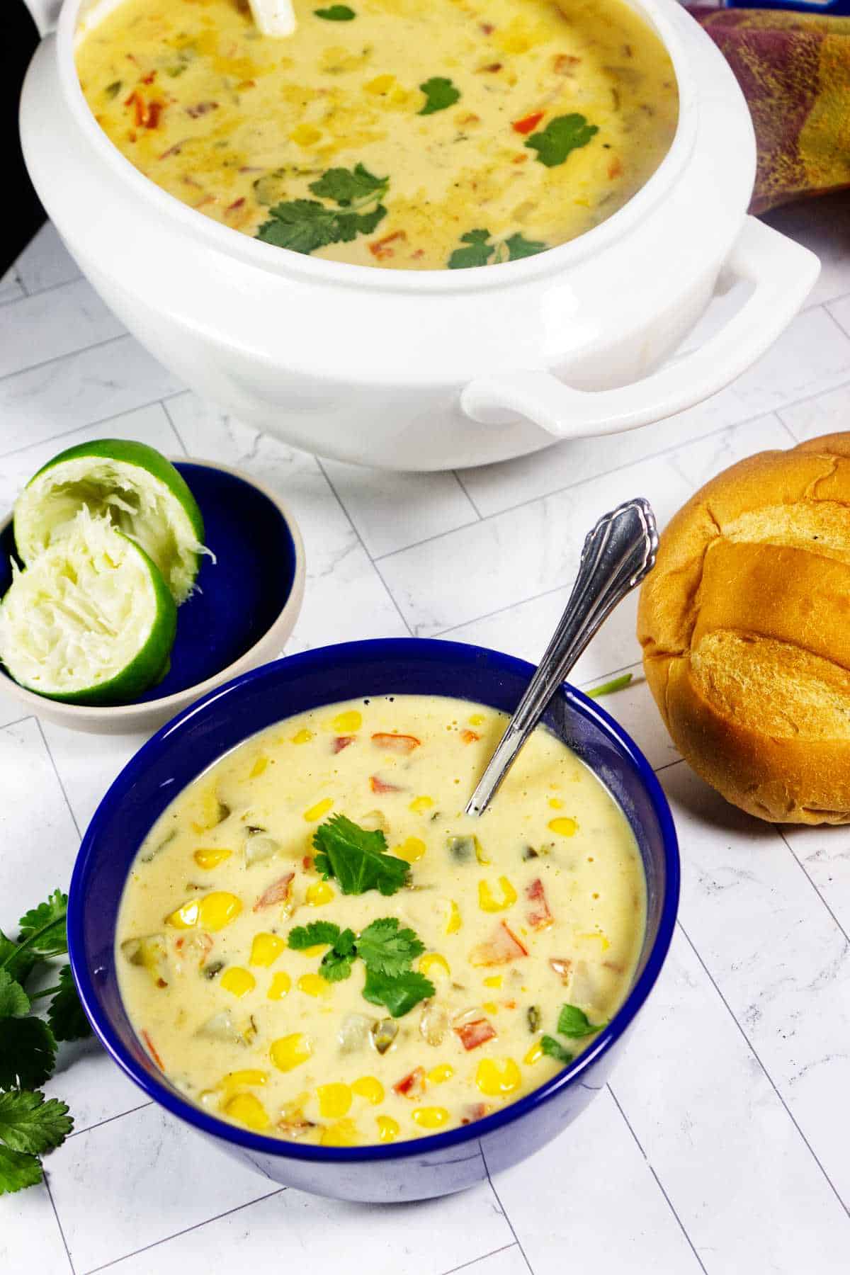 Panera Summer Corn Chowder in a blue bowl with bread, lime, cilantro, and soup tureen on a tiled surface.