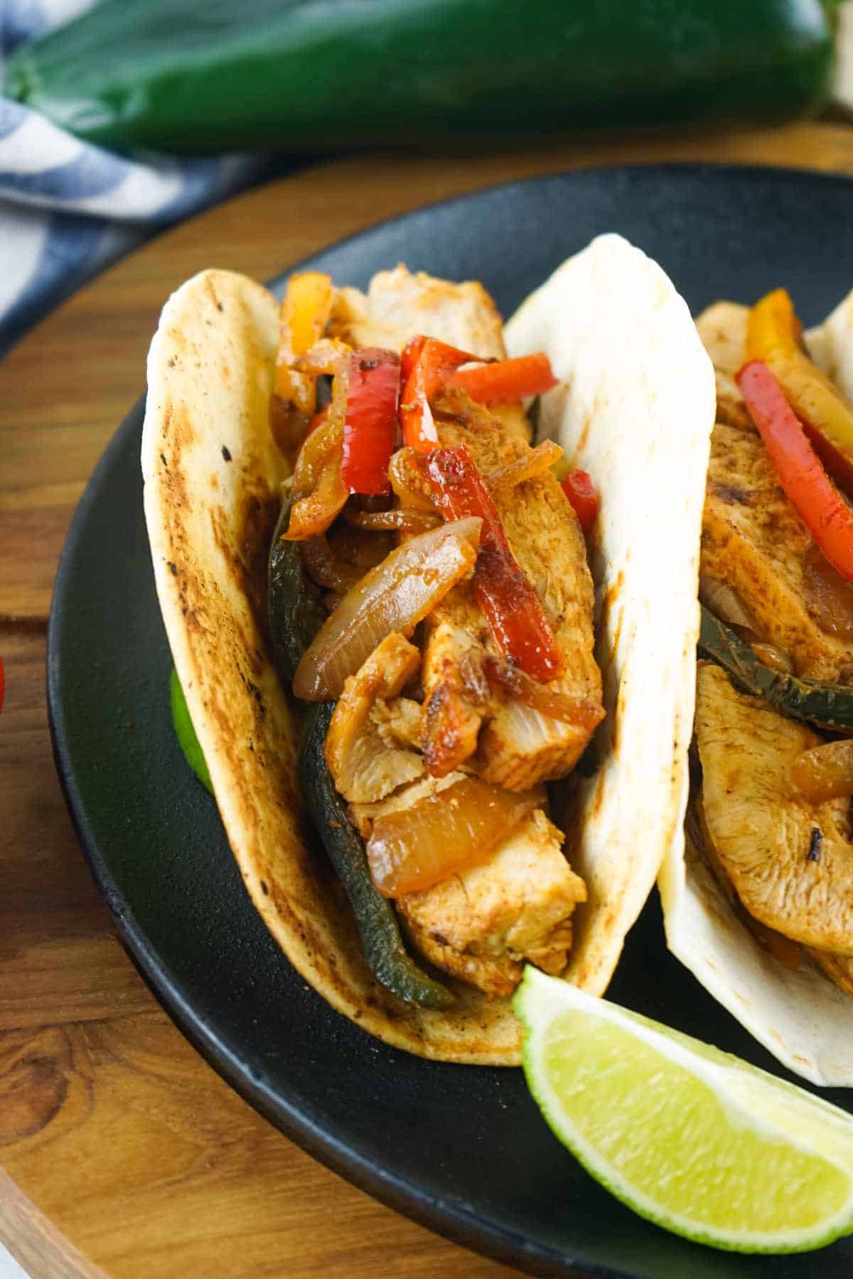 Chicken fajita tortilla with grilled chicken, peppers, onions, and lime on a black plate.