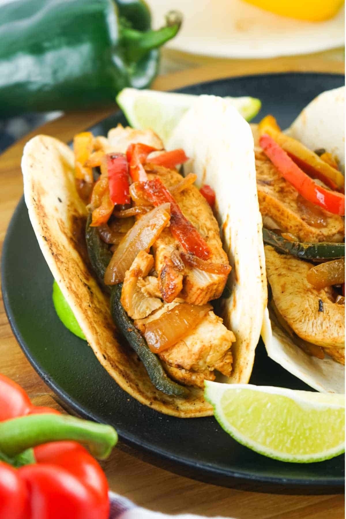 Chicken fajitas in tortillas with peppers, onions, lime wedges, and fresh peppers.