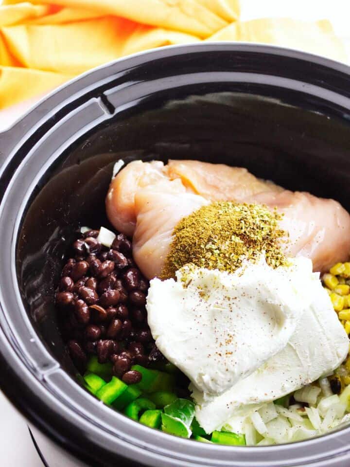 Slow cooker with chicken, black beans, corn, onion, cream cheese, and seasonings for chicken enchilada soup.