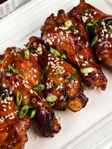 Four shiny, caramelized chicken drumsticks on a white plate, topped with sesame seeds and chopped green onions.