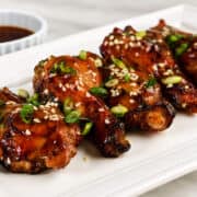 Four soy garlic chicken wings with sesame seeds and green onions on a white plate; brown dipping sauce in the background.