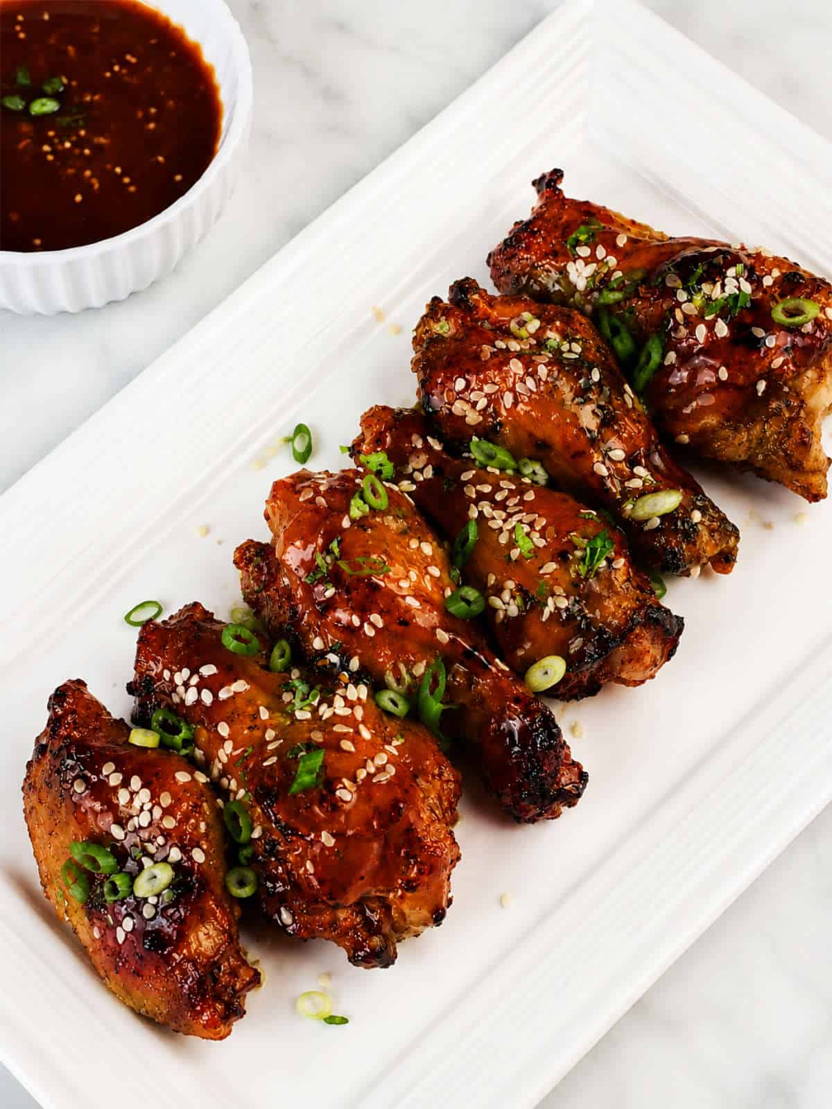 Soy garlic chicken wings with sesame seeds, sliced green onions, and dipping sauce on a white platter.