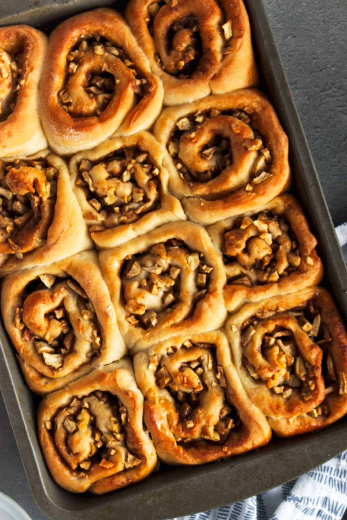 A pan of twelve golden brown cinnamon rolls with chopped nuts or apples on top rests on a dark surface.