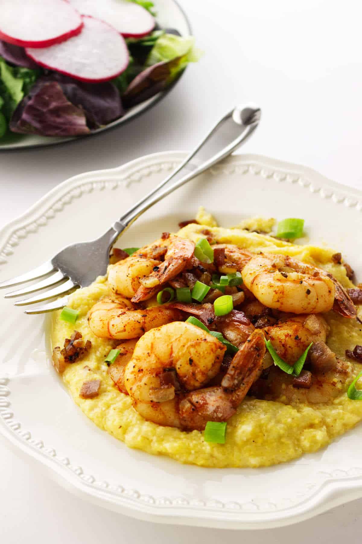 Shrimp and grits with green onions on a plate, fork nearby, and a leafy salad with radishes in the background.