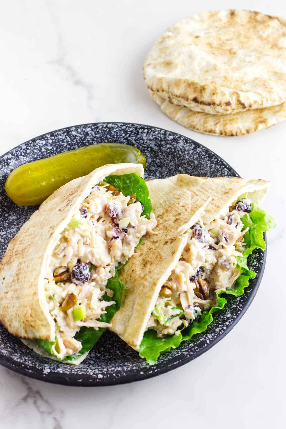 Two chicken salad pita pockets with lettuce and raisins on a speckled plate with a pickle; extra pita bread in the background.