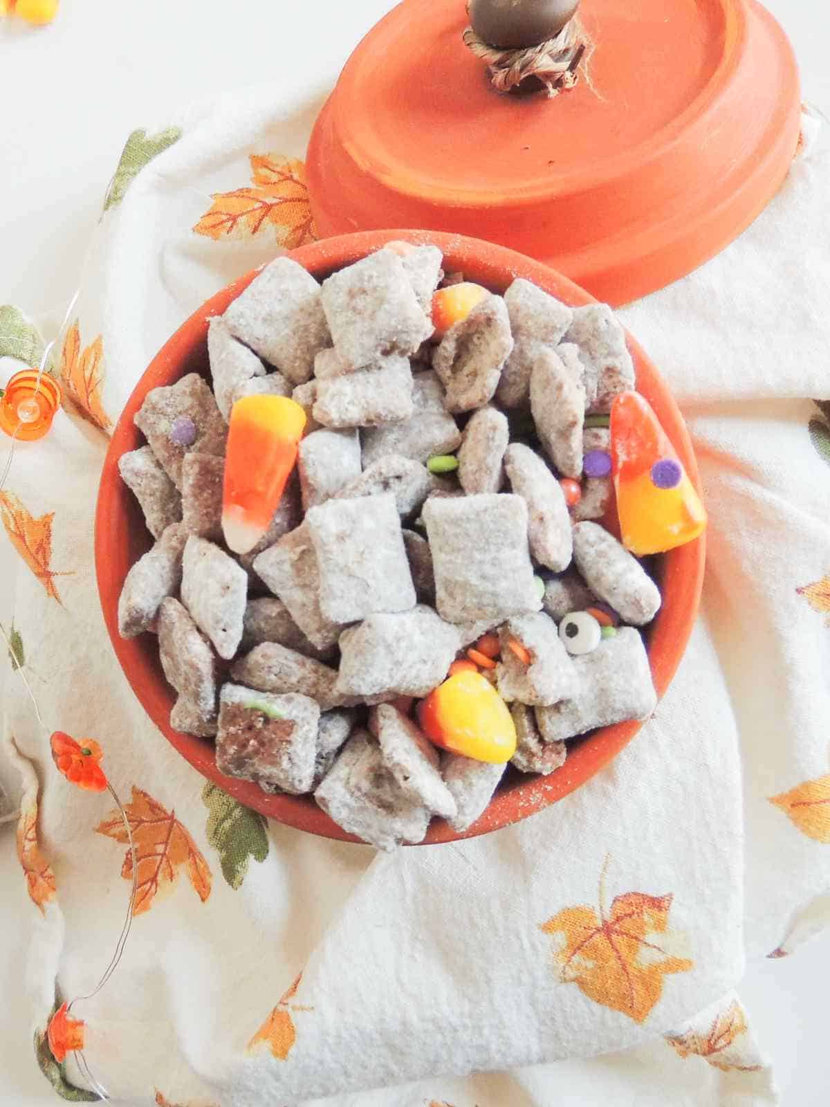 Snack mix coated in powdered sugar with candy corn and colorful candies in a bowl on an autumn leaf cloth, orange lid beside it.