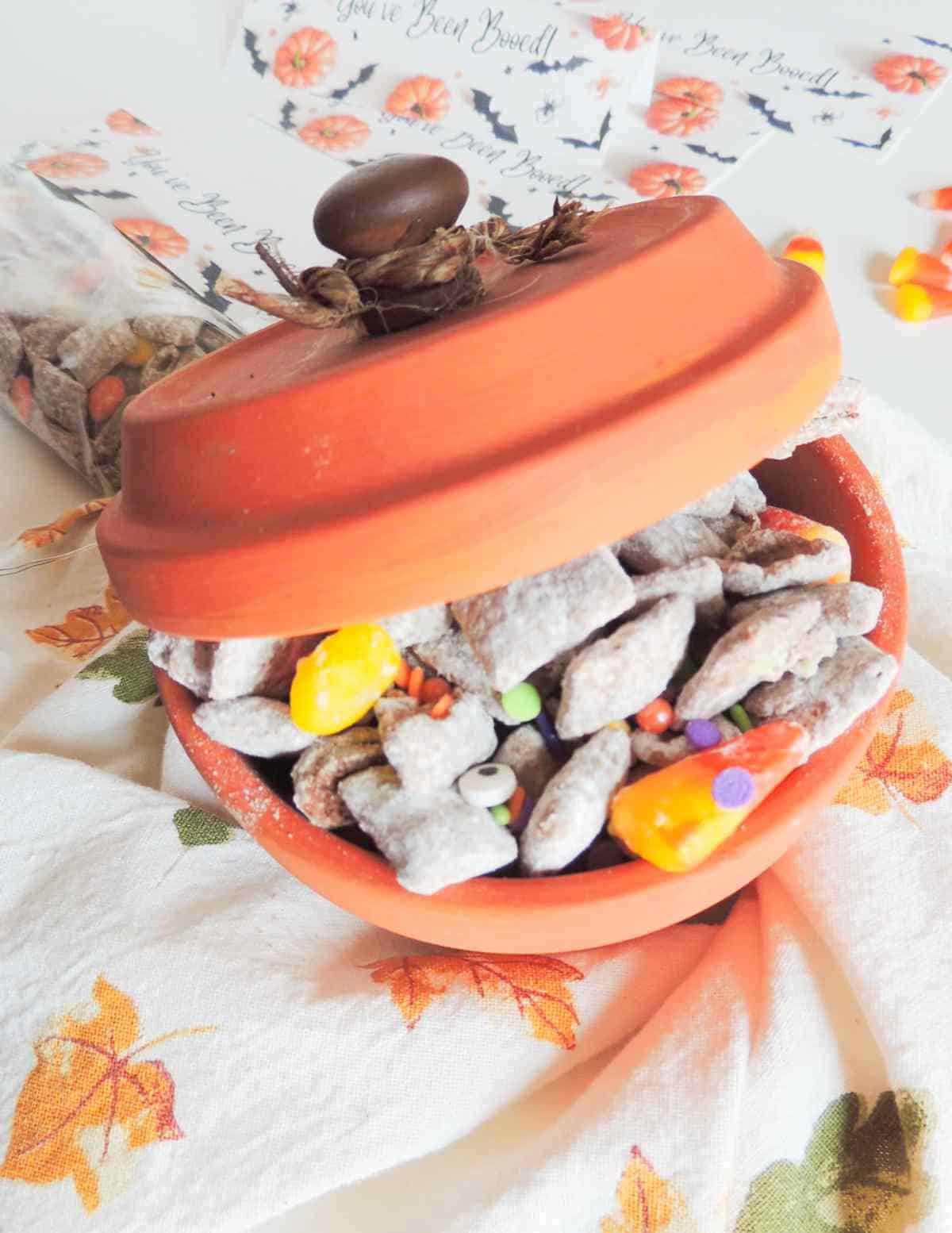 Terra cotta pot with powdered snack mix, candy corn, and colorful candies on a fall leaf napkin; lid slightly open.