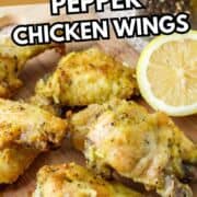 Honey lemon pepper chicken wings on a wooden board with black pepper, halved lemon, and a small bowl of yellow dipping sauce in the background.