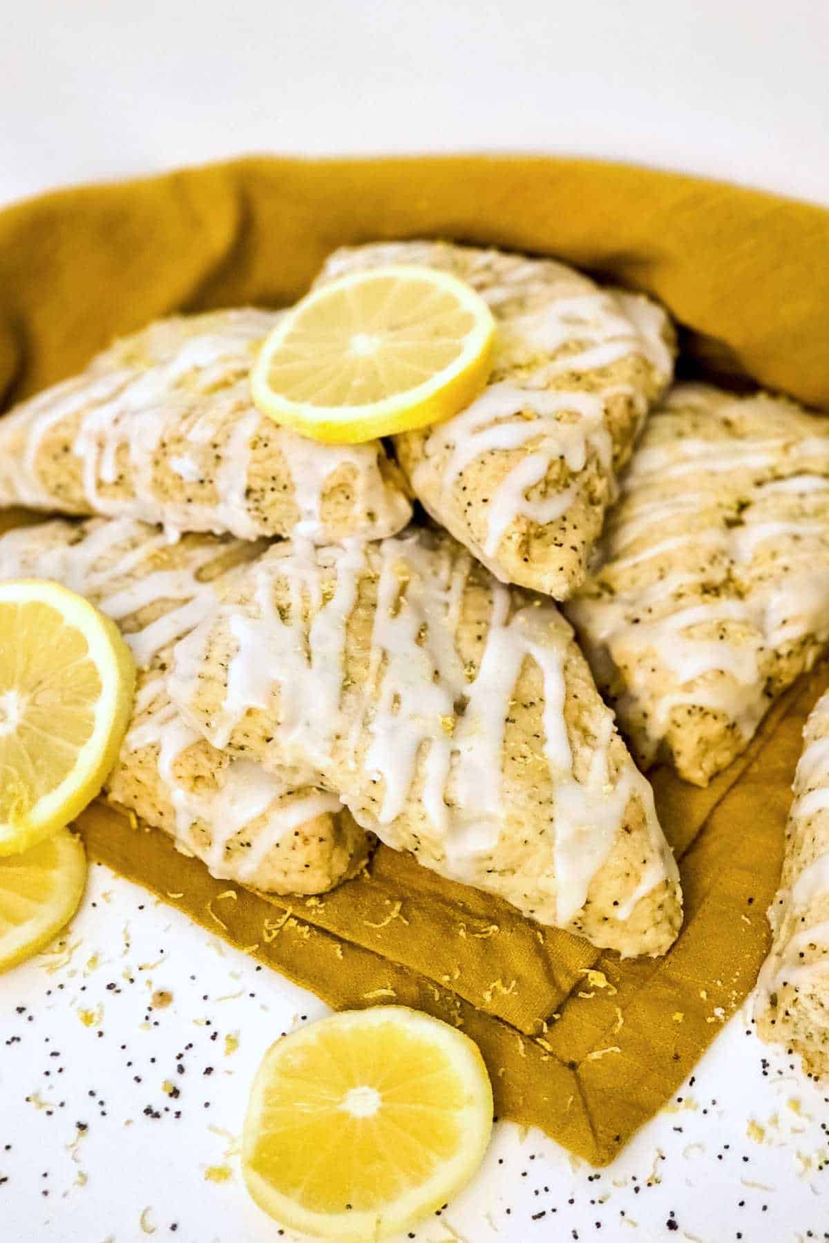 Lemon poppy seed scones with white icing stacked on a yellow cloth, topped with lemon slices and zest.