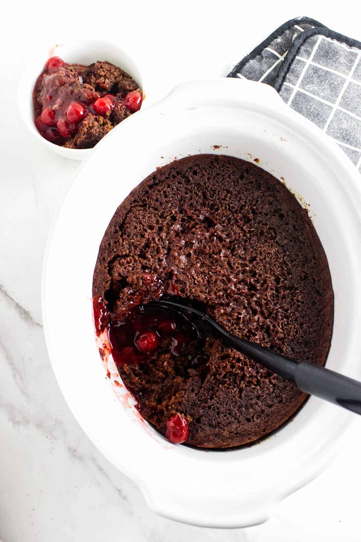 Chocolate cherry dump cake in a white slow cooker with a serving spoon and some scooped out. A small bowl of the dessert sits nearby on a white countertop next to a checkered oven mitt.