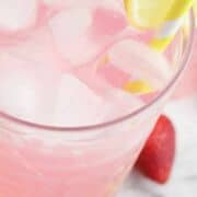 A glass of pink strawberry lemonade spritzer with ice, a lemon slice, and a yellow-striped straw. Another glass and a strawberry are in the background. Bottom text: "STRAWBERRY LEMONADE SPRITZER.