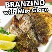 Baked branzino fillet with crispy skin, topped with golden brown miso glaze, served on a white plate with lime wedges and fresh herbs.