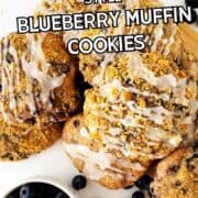Blueberry muffin cookies with icing on a plate, fresh blueberries around. Text: Crumbl style blueberry muffin cookies.
