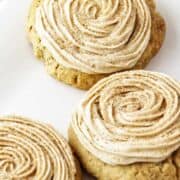 Three churro cookies with cinnamon-sugar swirled frosting sit on a white rectangular plate next to a label reading “CRUMBL CHURRO COOKIES.”.