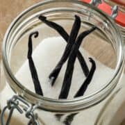 Four vanilla beans on white sugar in an open glass jar with a metal latch, top view.