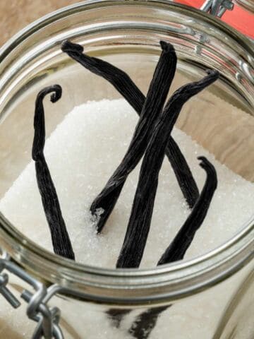 Four vanilla beans on white sugar in an open glass jar with a metal latch, top view.
