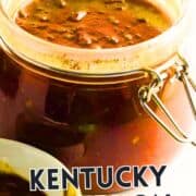 Glass jar of chunky brown Kentucky bourbon BBQ sauce with metal clasp, partially open on a white surface; some sauce in a nearby white dish. Text overlay: “Kentucky Bourbon BBQ Sauce.”.