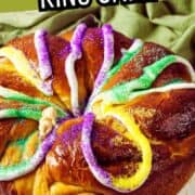 Round king cake with purple, green, and yellow sugar and icing on a white plate; “Mardi Gras King Cake” banner overhead.
